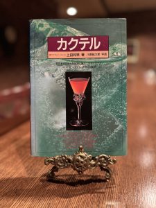 バーテンダーを志すきっかけとなったカクテルブック。木製のカウンターの上に飾られ、表紙には鮮やかなカクテルの写真と『カクテル』のタイトルが記されている。