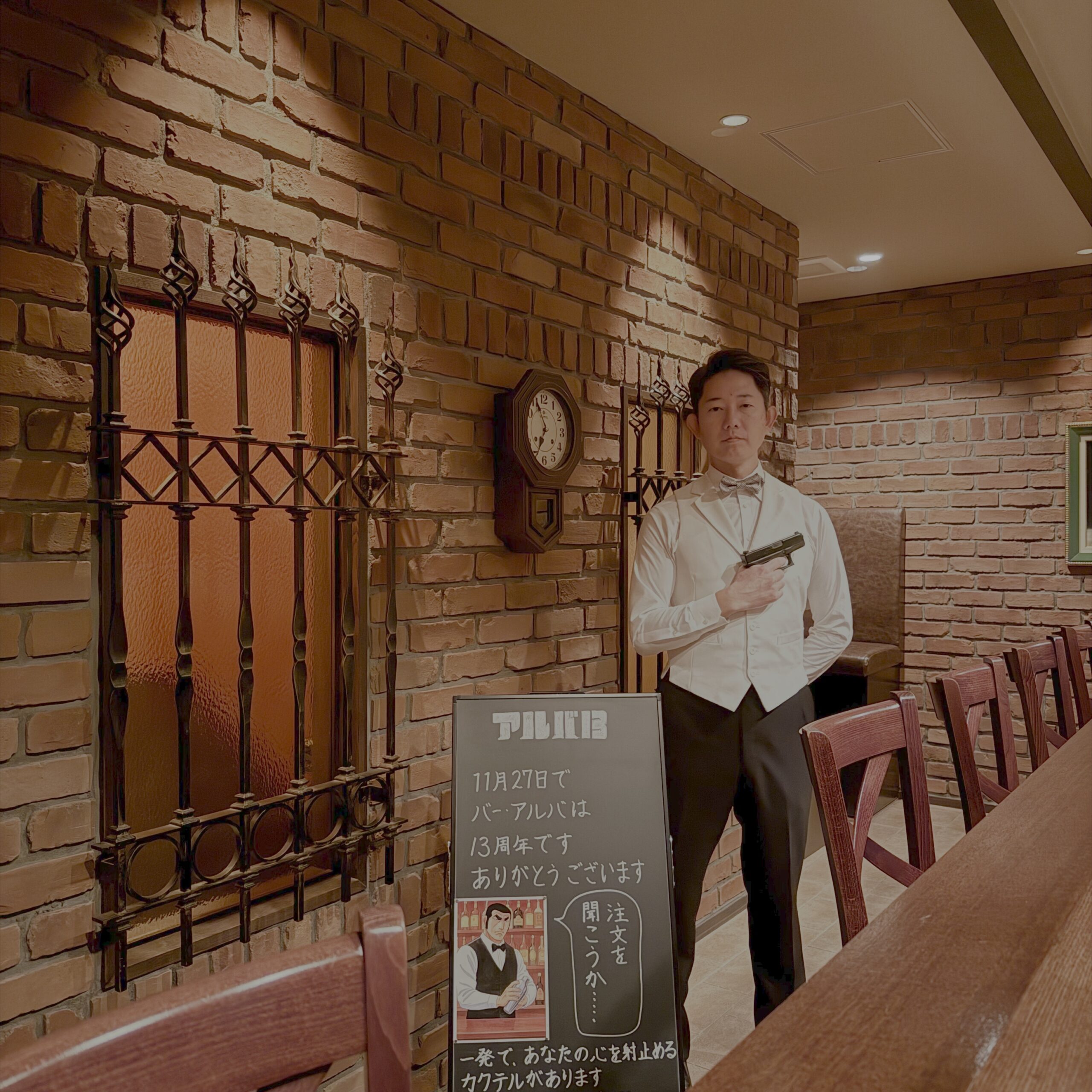 BAR ALBAの店内で、13周年を記念した「アルバ13」黒板の前に立つバーテンダーの写真。天満・天神橋の落ち着いたレンガ造りのバーカウンター。